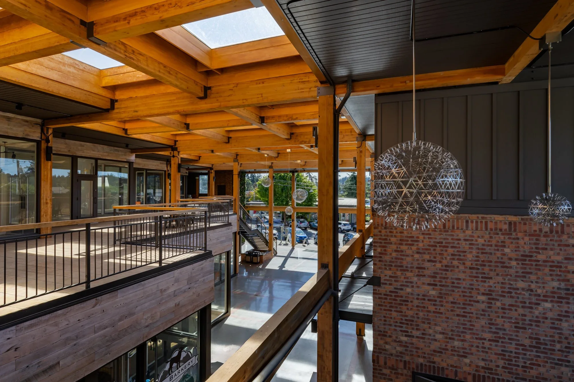 Interior of a modern building with wooden beams, large skylights, and hanging spherical light fixtures, overlooking a parking lot outside.