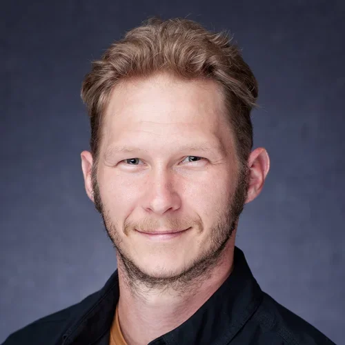 A portrait of a man with short, light brown hair and a beard, wearing a dark shirt, against a plain dark background.