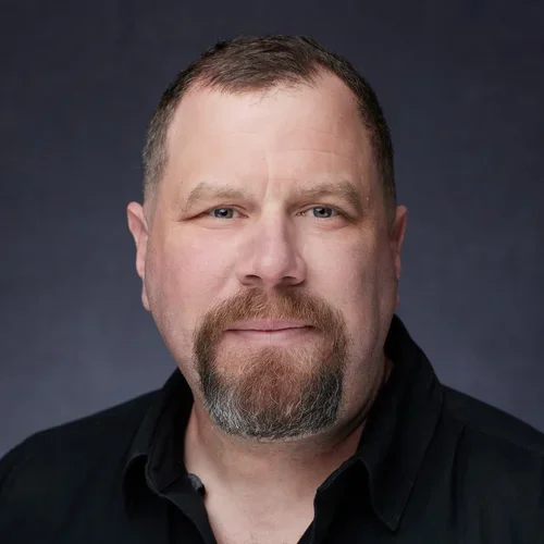 Close-up portrait of a man with short brown hair, a beard, and mustache, wearing a black shirt, against a dark background.