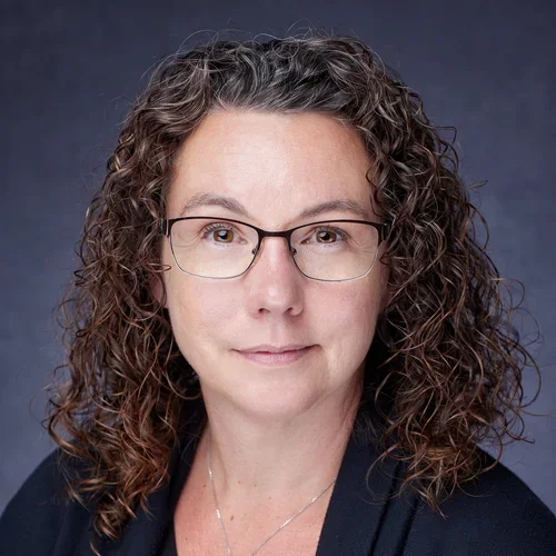 A woman with curly brown hair and glasses, wearing a dark blazer, looking directly at the camera against a dark background.