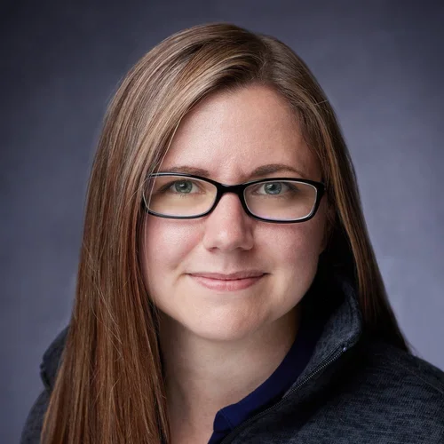 A woman with long brown hair, glasses, and a slight smile wearing a dark jacket against a gray background.