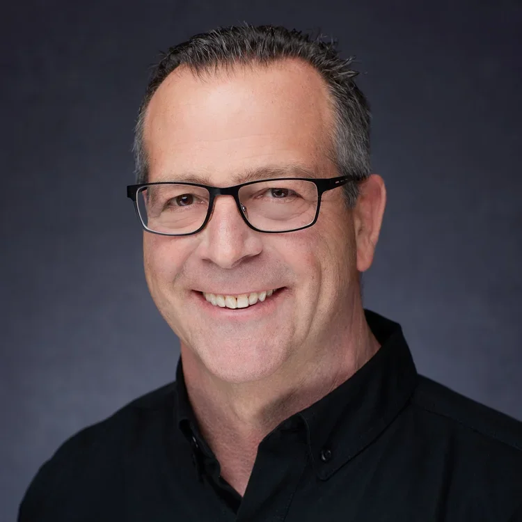 Headshot of a middle-aged man with short, gray hair wearing glasses and a black collared shirt, smiling against a dark background.