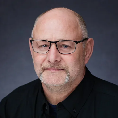 Portrait of a middle-aged man with glasses, a beard, and a mustache, wearing a black shirt against a dark background.