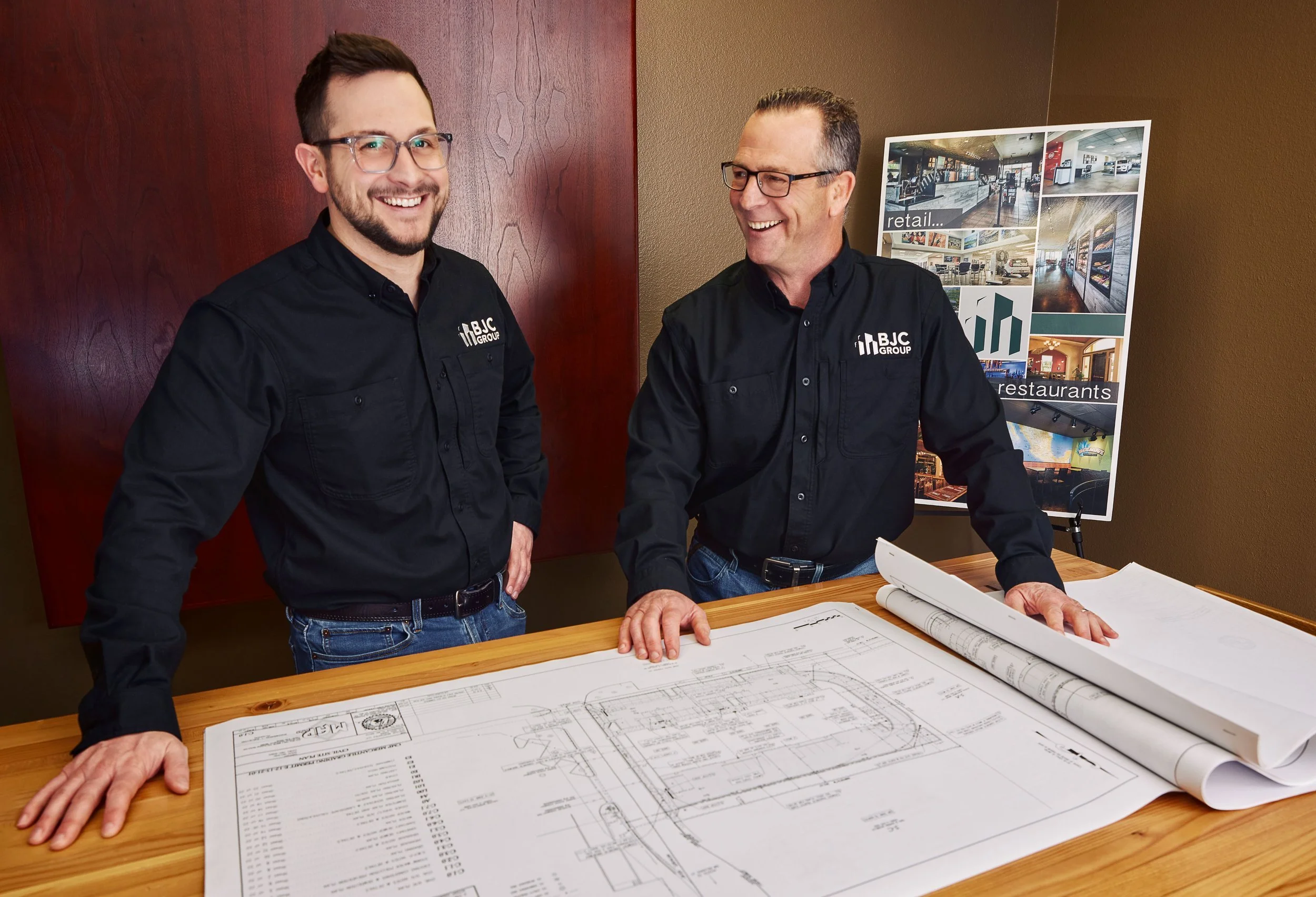 Two men in black shirts with BJC Group logo standing at a wooden table with architectural blueprints. They are smiling and engaging in conversation. Behind them is a wall with a display of interior photos and the words 'retail' and 'restaurants'.