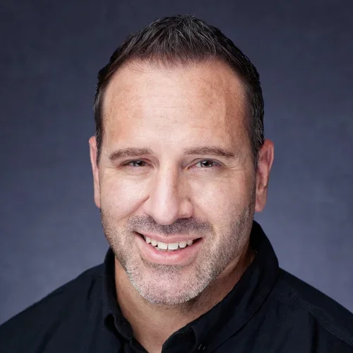 Headshot of a smiling man with short dark hair, wearing a black shirt, against a dark background.