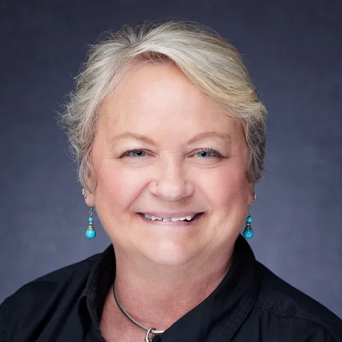 A smiling middle-aged woman with short blonde hair, wearing turquoise earrings, a black top, and a silver necklace, against a dark background.