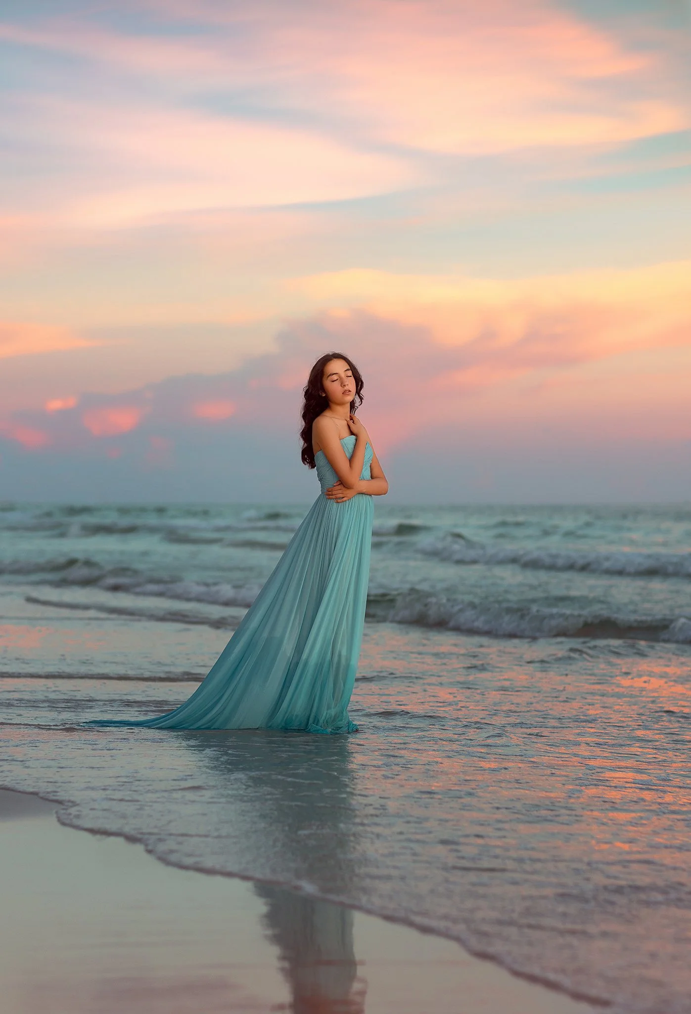 A woman in a long turquoise dress standing in the shallow surf at the beach during sunset, with her eyes closed and arms crossed over her chest.