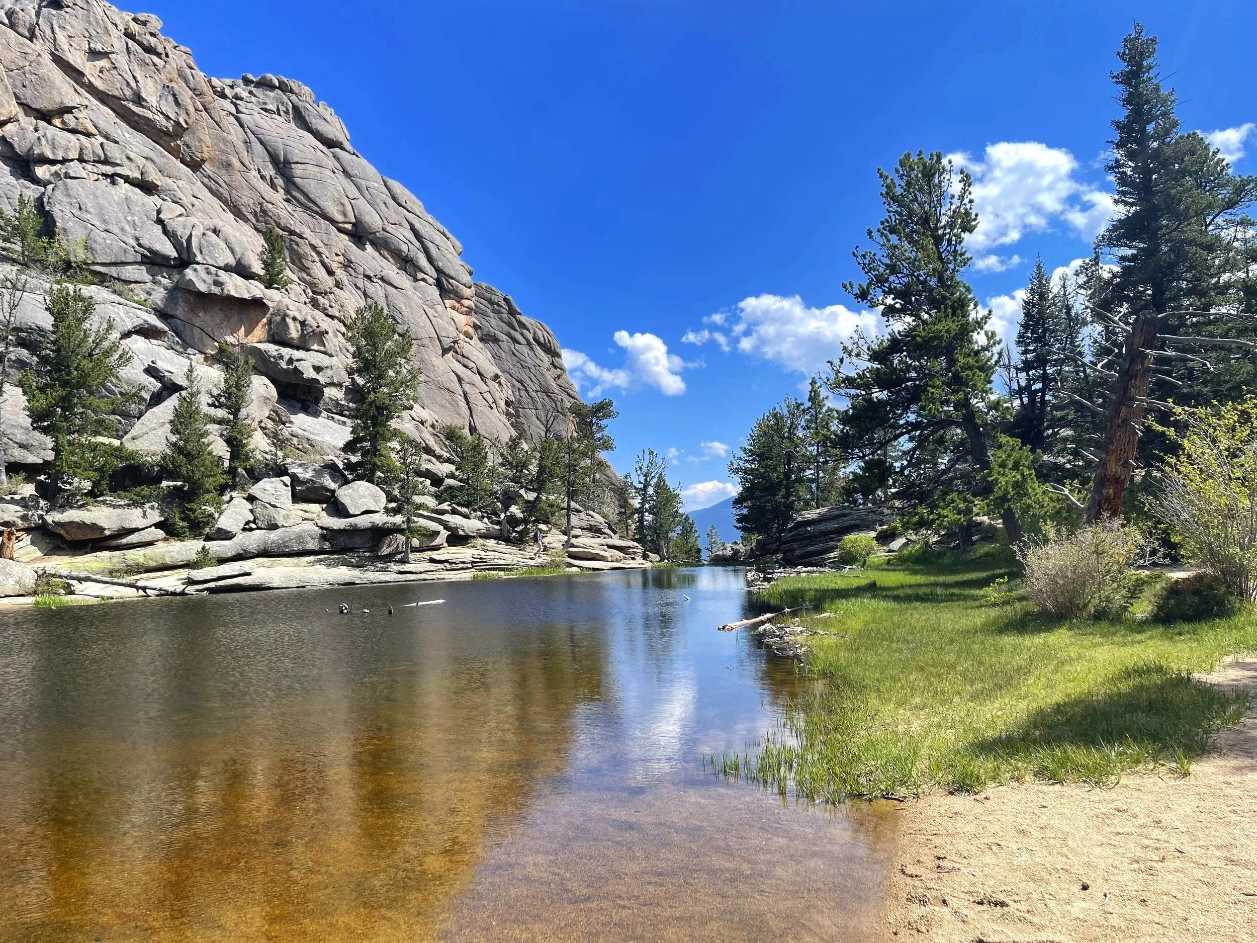 A serene lakeside scene with clear water reflecting the blue sky, surrounded by tall green pine trees and rocky cliffs in the background, under a mostly sunny sky with scattered white clouds.