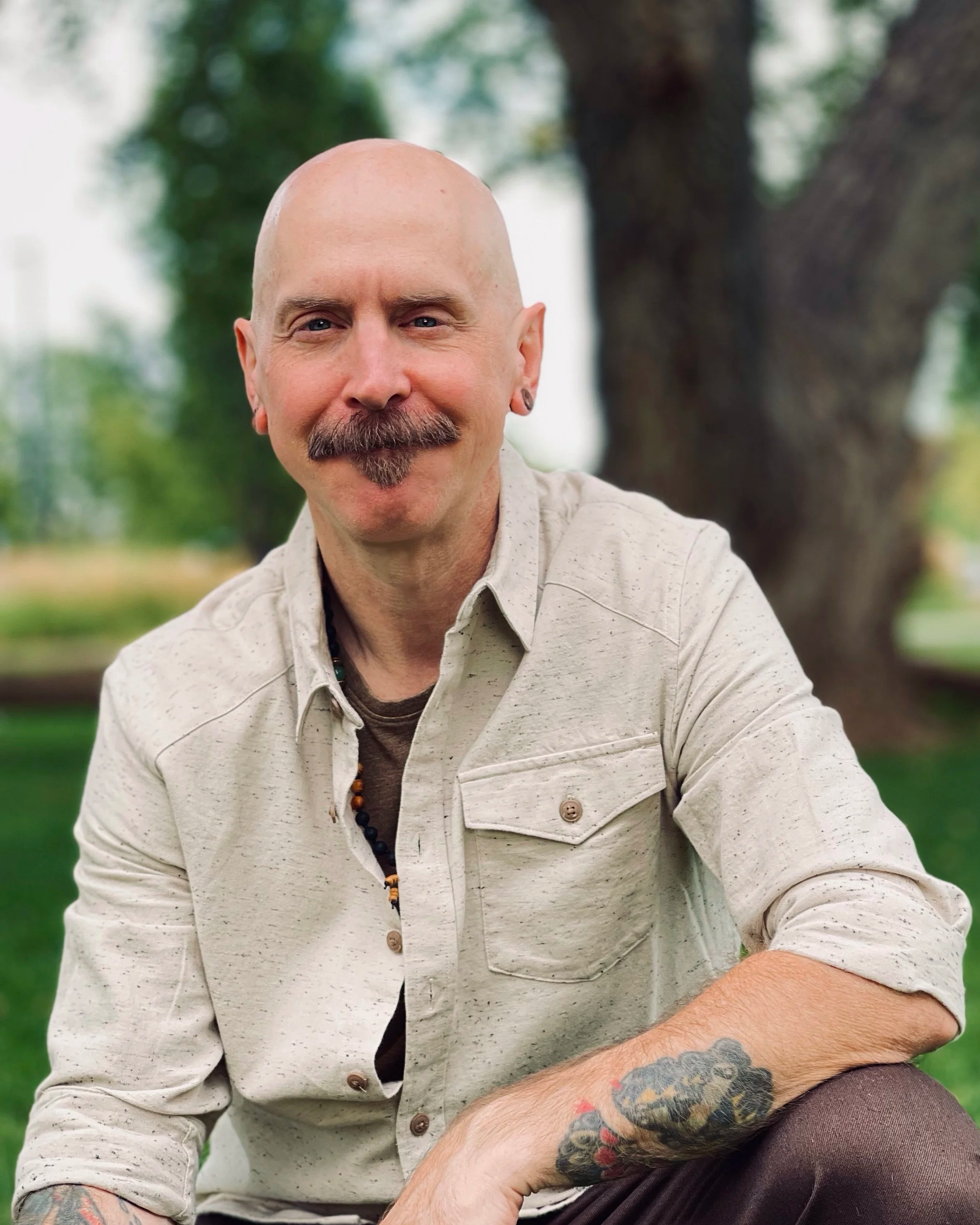 A bald man with a mustache and beard, wearing a beige shirt with rolled-up sleeves, sits outdoors in front of a large tree and green grass, smiling at the camera.