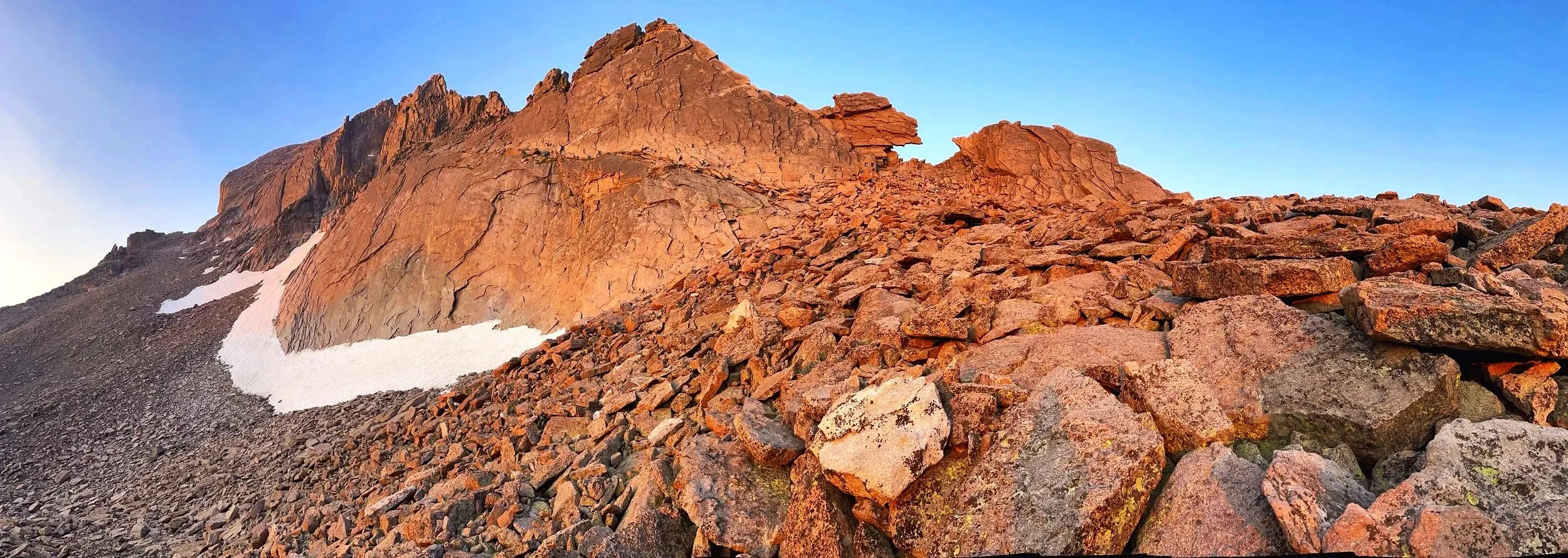 A rugged mountain landscape with reddish-brown rocks and a small patch of snow, under a clear blue sky.