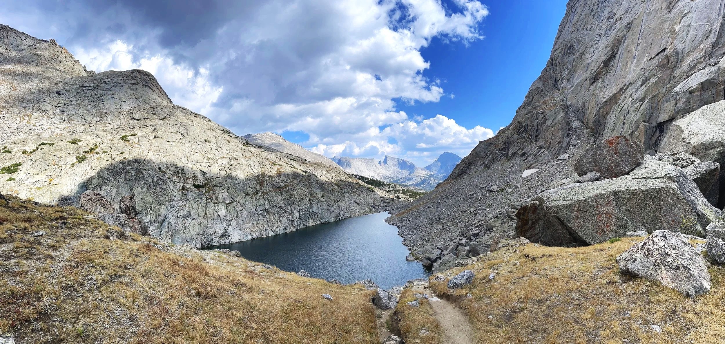 A mountain landscape with a narrow lake at the base of rocky slopes, under a partly cloudy sky.
