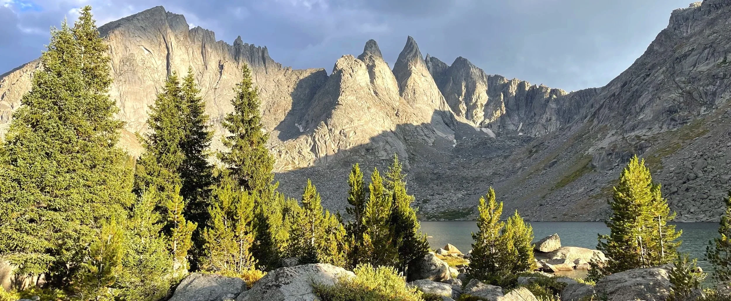 Mountains with rocky peaks, green trees in the foreground, a lake, and a partly cloudy sky