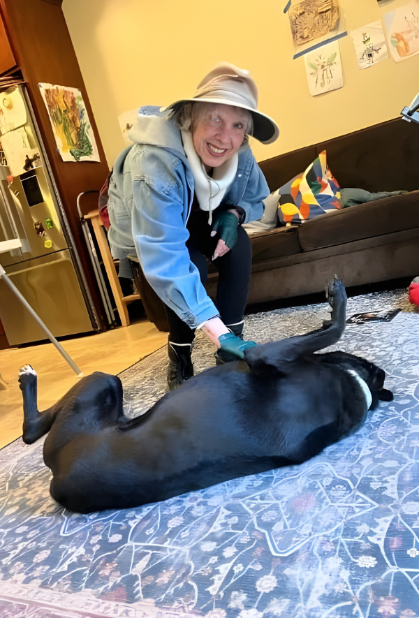 BB, positive reinforcement dog trainer in Oakland, CA, wears a wide-brimmed hat and is smiling as she interacts with a large black dog lying on its back on a patterned rug inside a room.