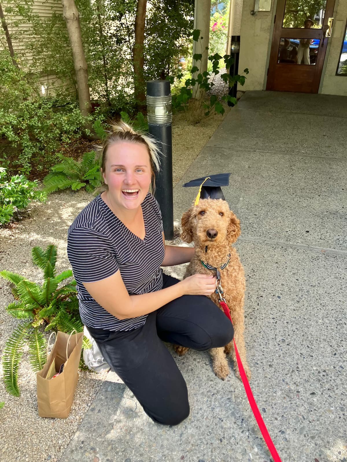 A woman smiling and squatting next to a dog wearing a graduation cap outside a building in Oakland, CA.