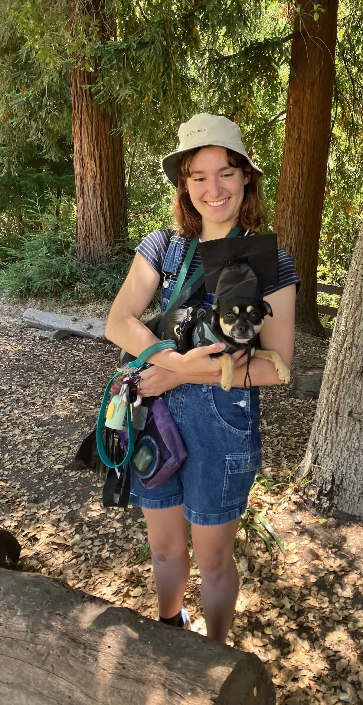 A young woman in a beige hat, striped shirt, and denim shorts holding a small black and tan dog wearing a graduation cap in a forested area in Oakland, CA.
