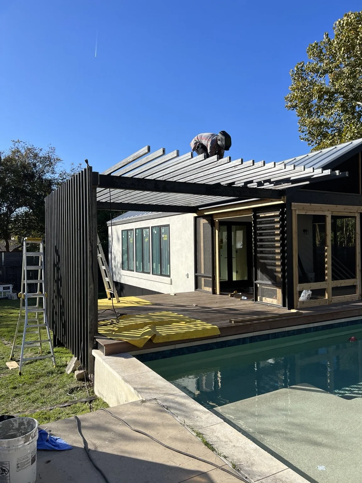 Metal pergola with overhead slats and a vertical screen being assembled beside a pool.