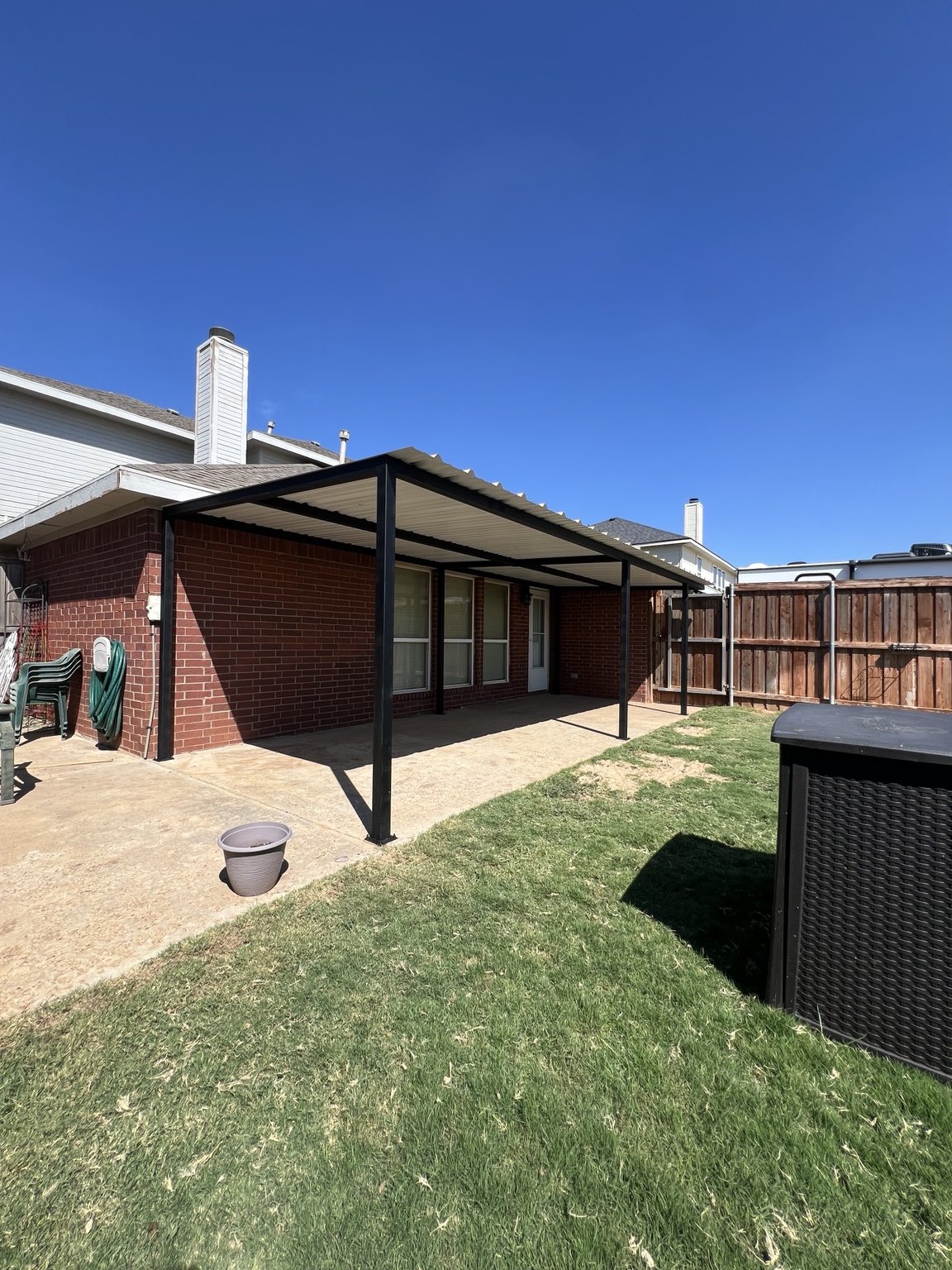 Black metal patio cover with light roof panels beside a brick house.