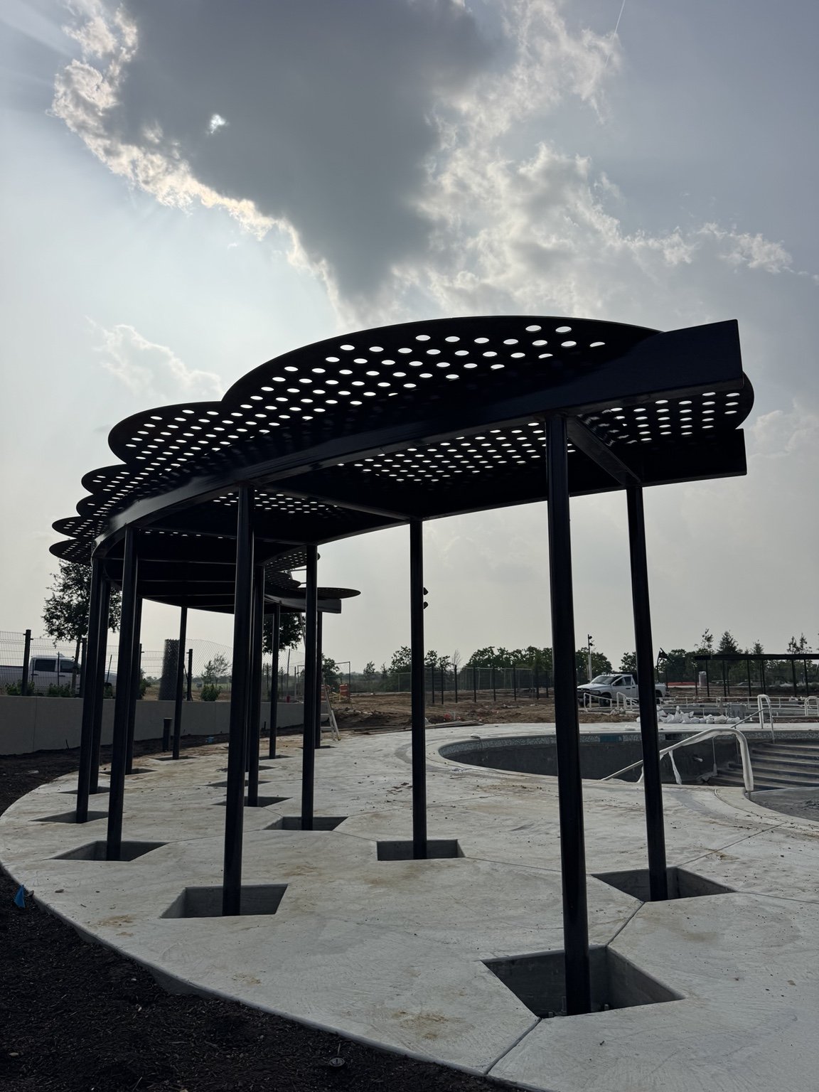 Curved metal shade structure with perforated roof panels beside a pool.