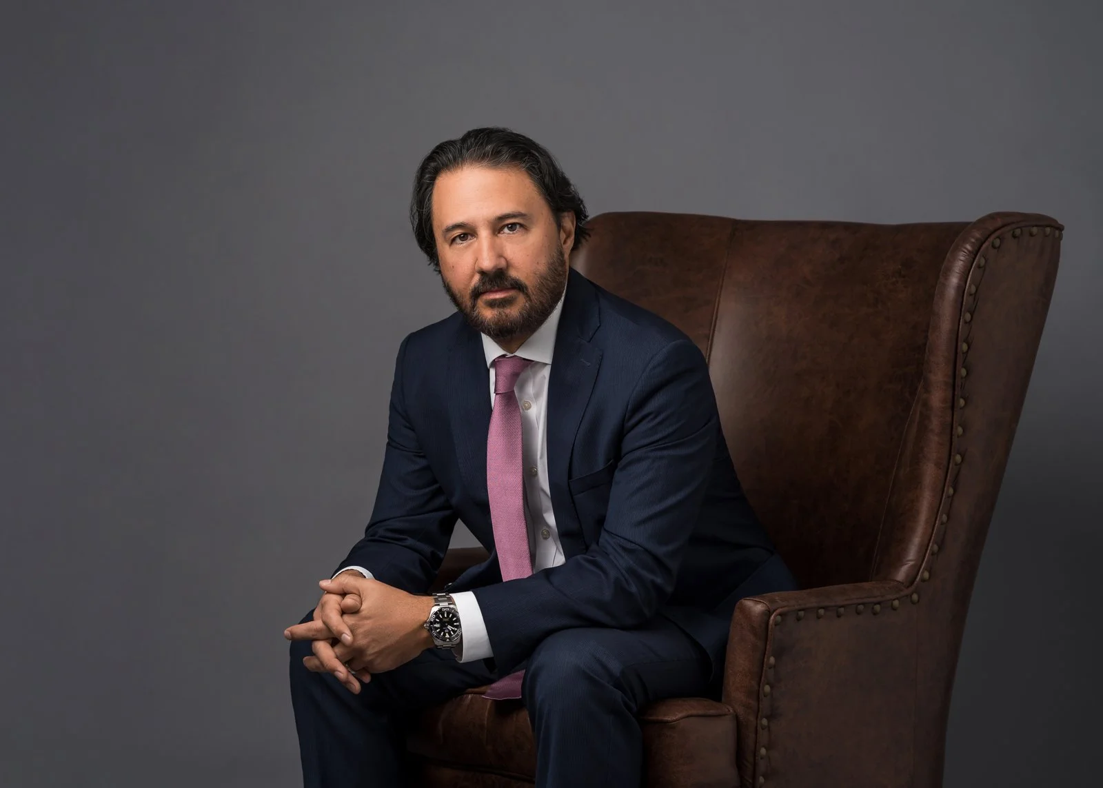 A man in a suit and tie seated on a chair, looking confidently at the camera.