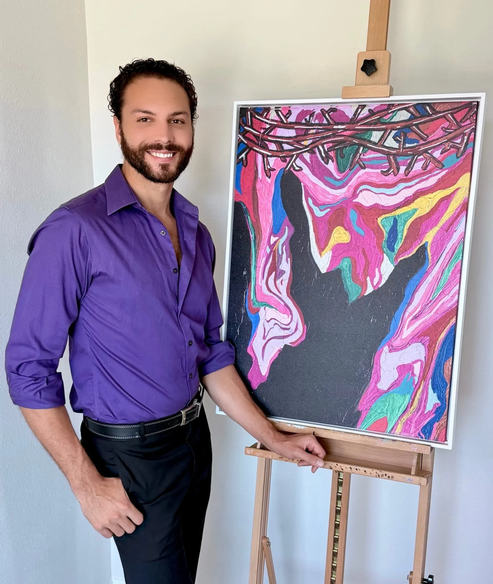A man with a beard and curly hair wearing a purple shirt, standing next to an abstract colorful painting on an easel, smiling at the camera.