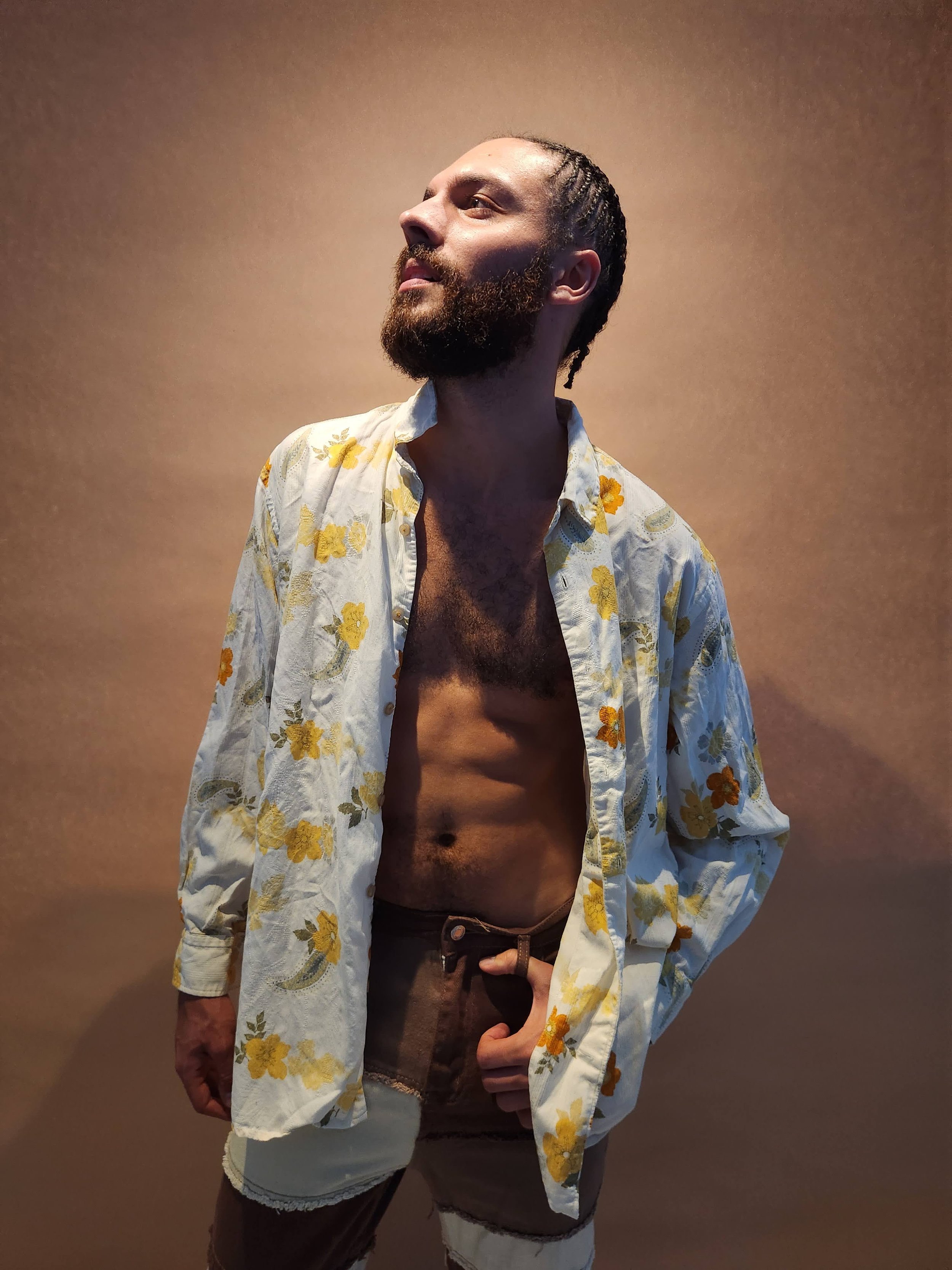 A man with a beard and braided hair wearing an open floral shirt and distressed shorts, standing against a plain background.