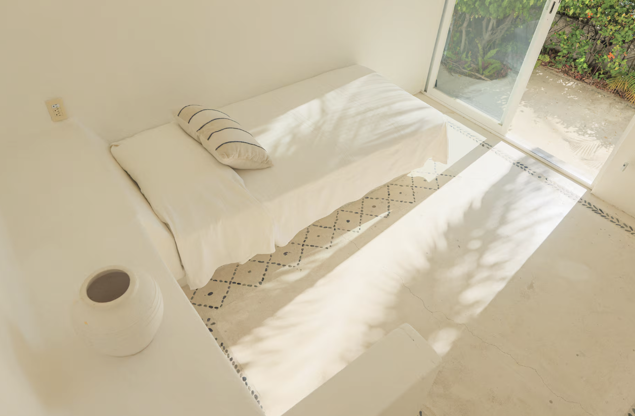 Bright room with white sofa, pillow, and vase, illuminated by natural sunlight through a sliding glass door, leading to a garden outside.