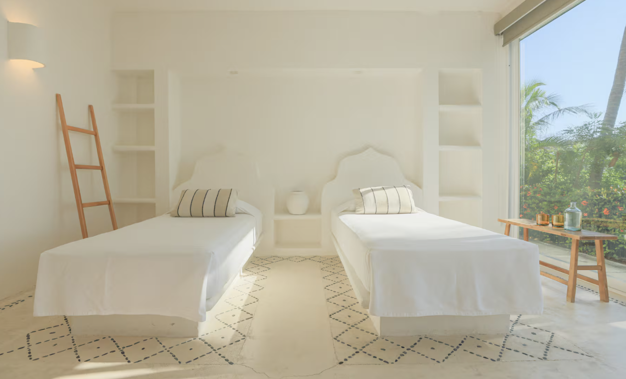 A bright, airy room with two single beds with white bedding, a bedside table with a vase, a wooden ladder, and a large window showing palm trees outside, decorated in a minimalist style.