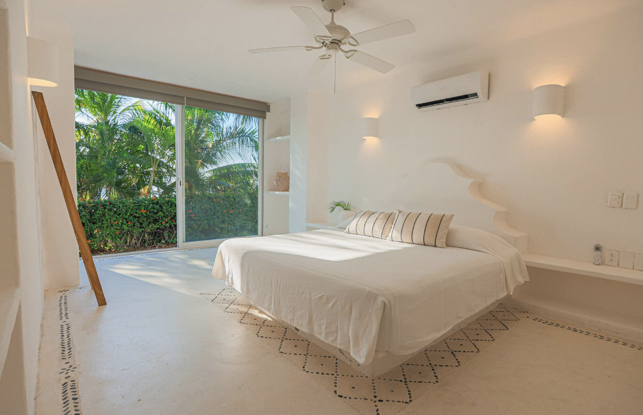 Bright hotel room with a large bed, white bedding, and striped pillows. Large sliding glass door shows green tropical foliage outside. The room has white walls, a ceiling fan, and minimal decor.