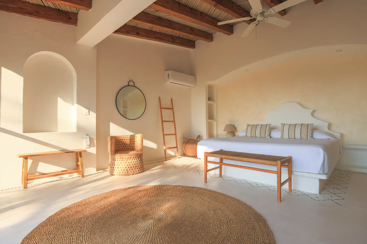 A bedroom with a large bed, woven bench, wicker basket, wooden ladder, small bench, round mirror, air conditioning, and ceiling fan, with natural sunlight illuminating the space.