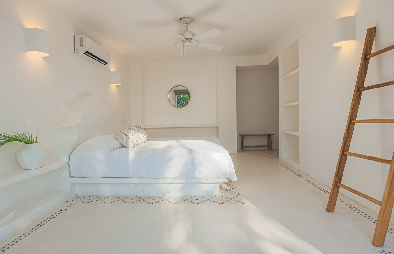 A minimalist bedroom with white walls, a white bed with striped pillow, a ceiling fan, a mirror, a wooden ladder, and built-in shelves.