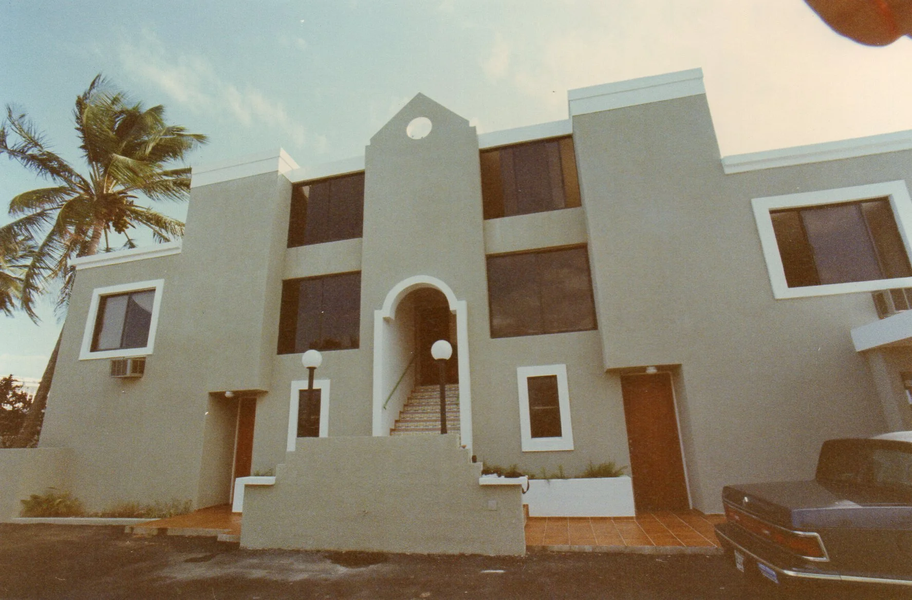 Edificio de estilo moderno con varias ventanas rectangulares grandes, entrada elevada con escaleras y luminarias en la fachada, palmera a la izquierda y coche oscuro estacionado en el frente.