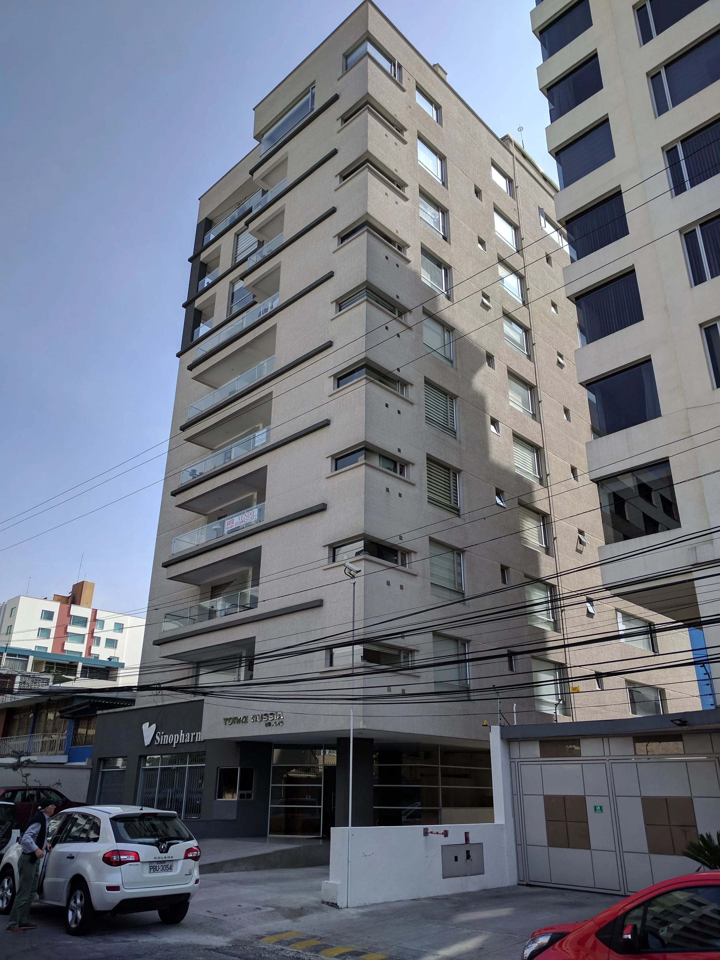 Edificio de apartamentos de varios pisos, con balcones y ventanas, en una calle urbana con autos estacionados y cables eléctricos.