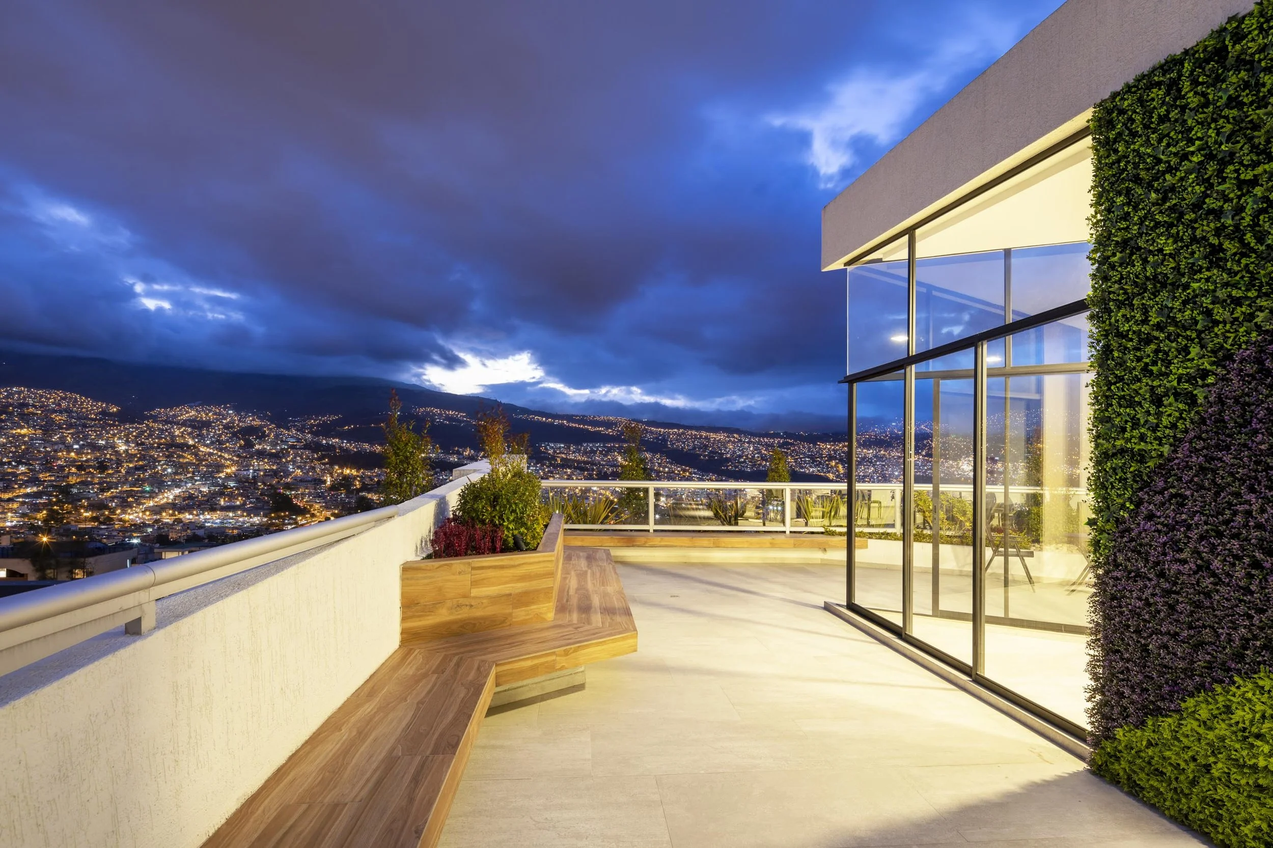 Vista de una terraza moderna con ventanales de cristal, plantas y ciudad iluminada al atardecer, cielo nublado.