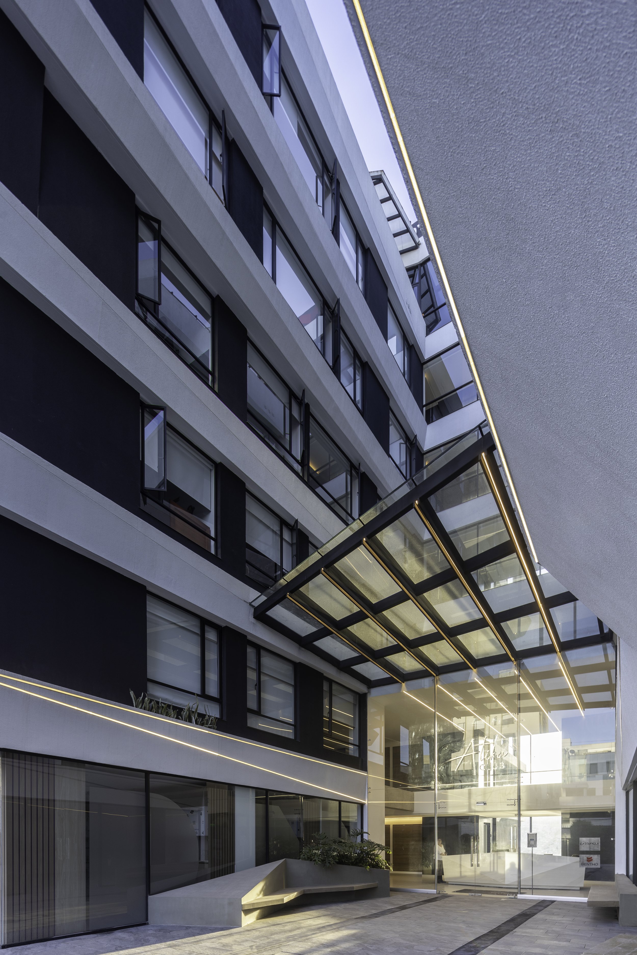 Vista de un edificio moderno con ventanas grandes y un área de entrada con estructura de vidrio y líneas de iluminación.