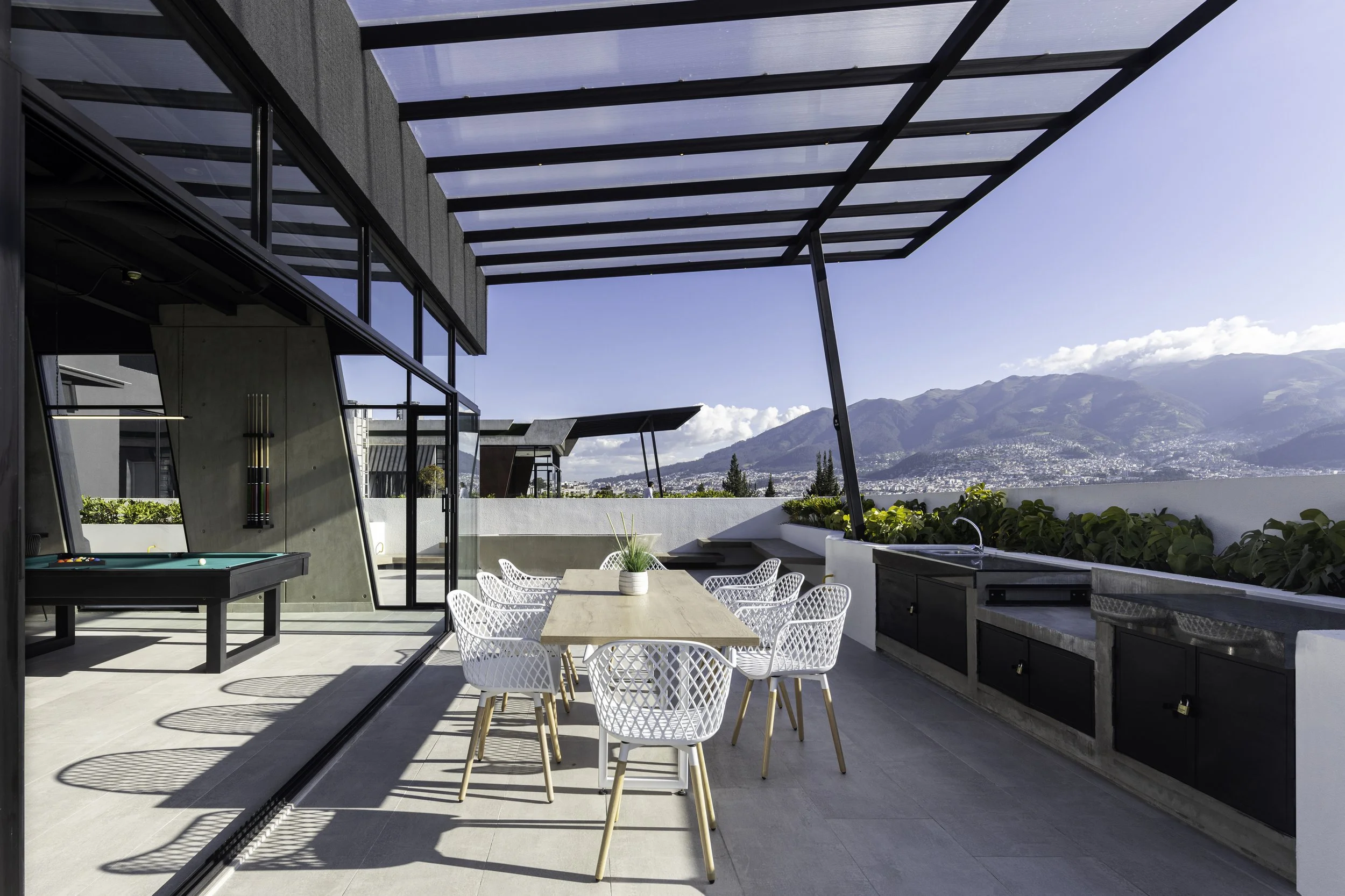 Terraza moderna con vista a las montañas, mesas y sillas blancas, área de cocina con fregadero y gabinetes oscuros, estructura de vidrio y metal, decoración sencilla y luminosa.