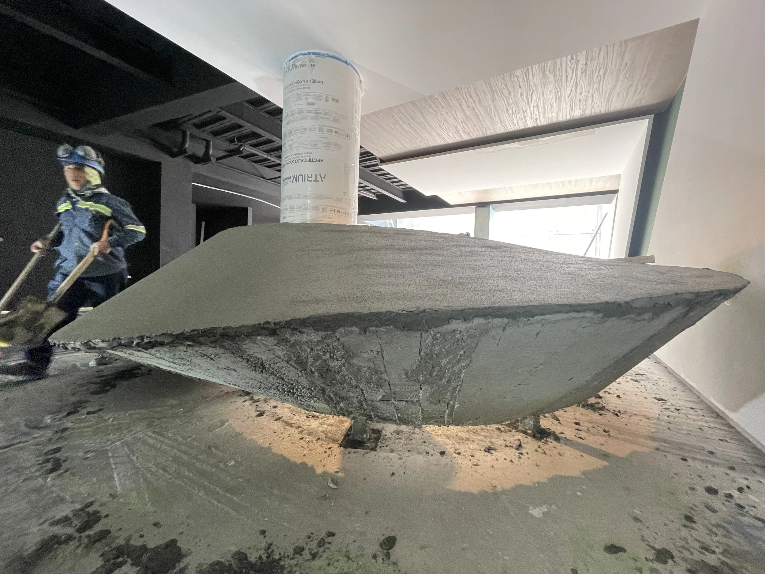 Interior de una construcción en proceso con una estructura de concreto en forma de embarcación, un cilindro de papel de construcción y una persona con equipo de protección laboral en el fondo.