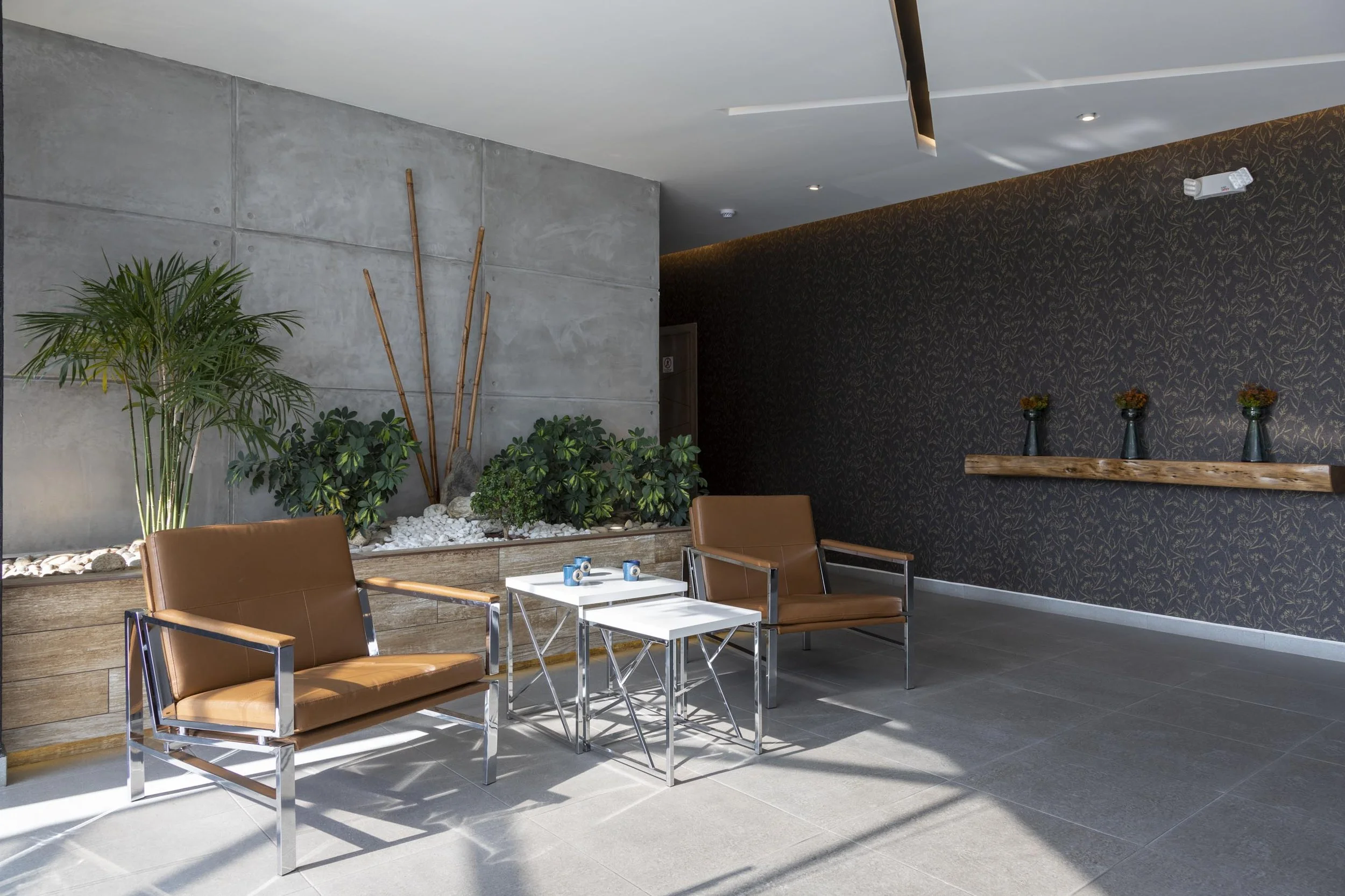 Área de descanso con sillas de cuero marrón, mesas blancas, plantas verdes y pared decorativa con flores en jarrones.