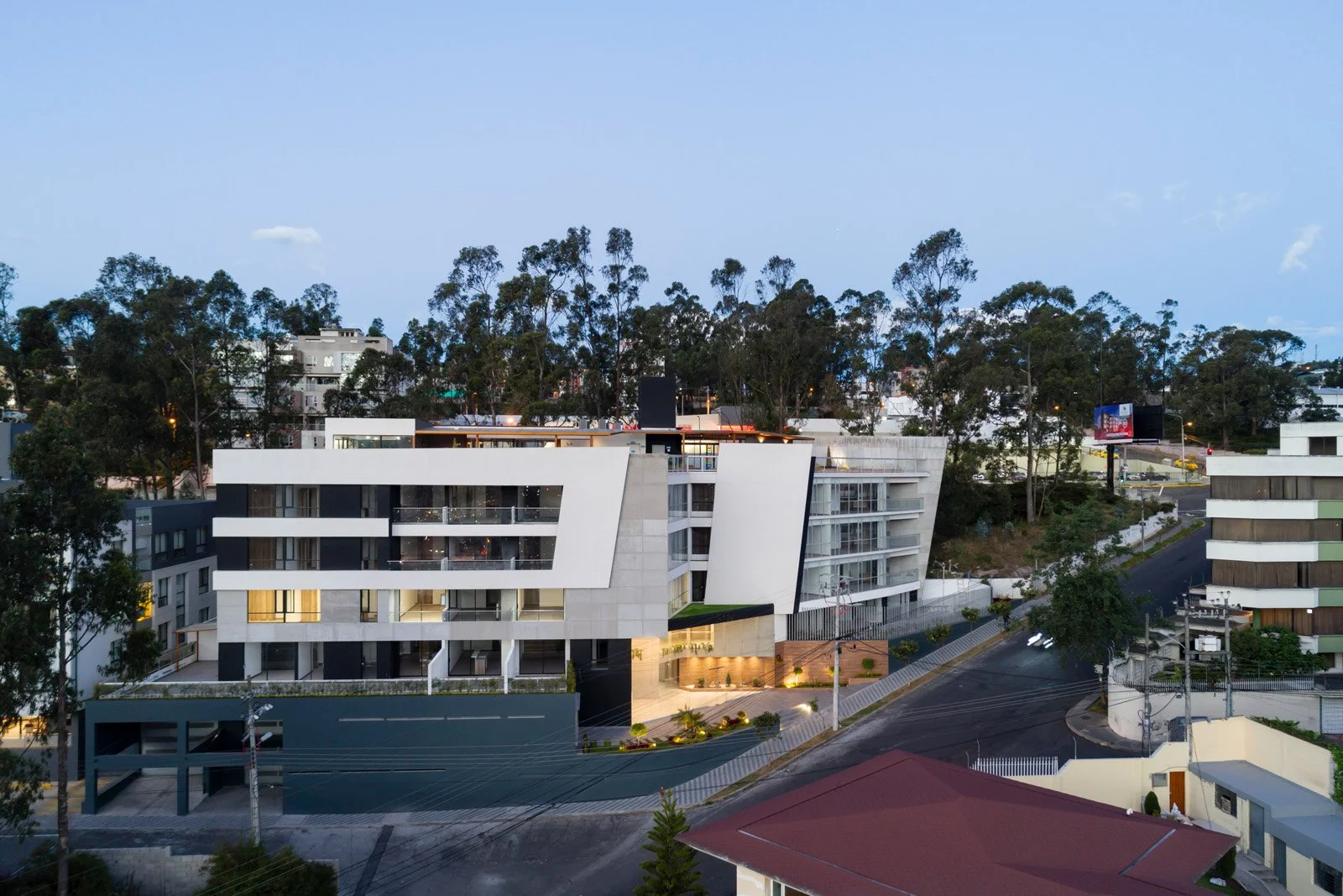 Edificio moderno de varios pisos con líneas geométricas, rodeado de árboles y calles en una zona urbana al atardecer.