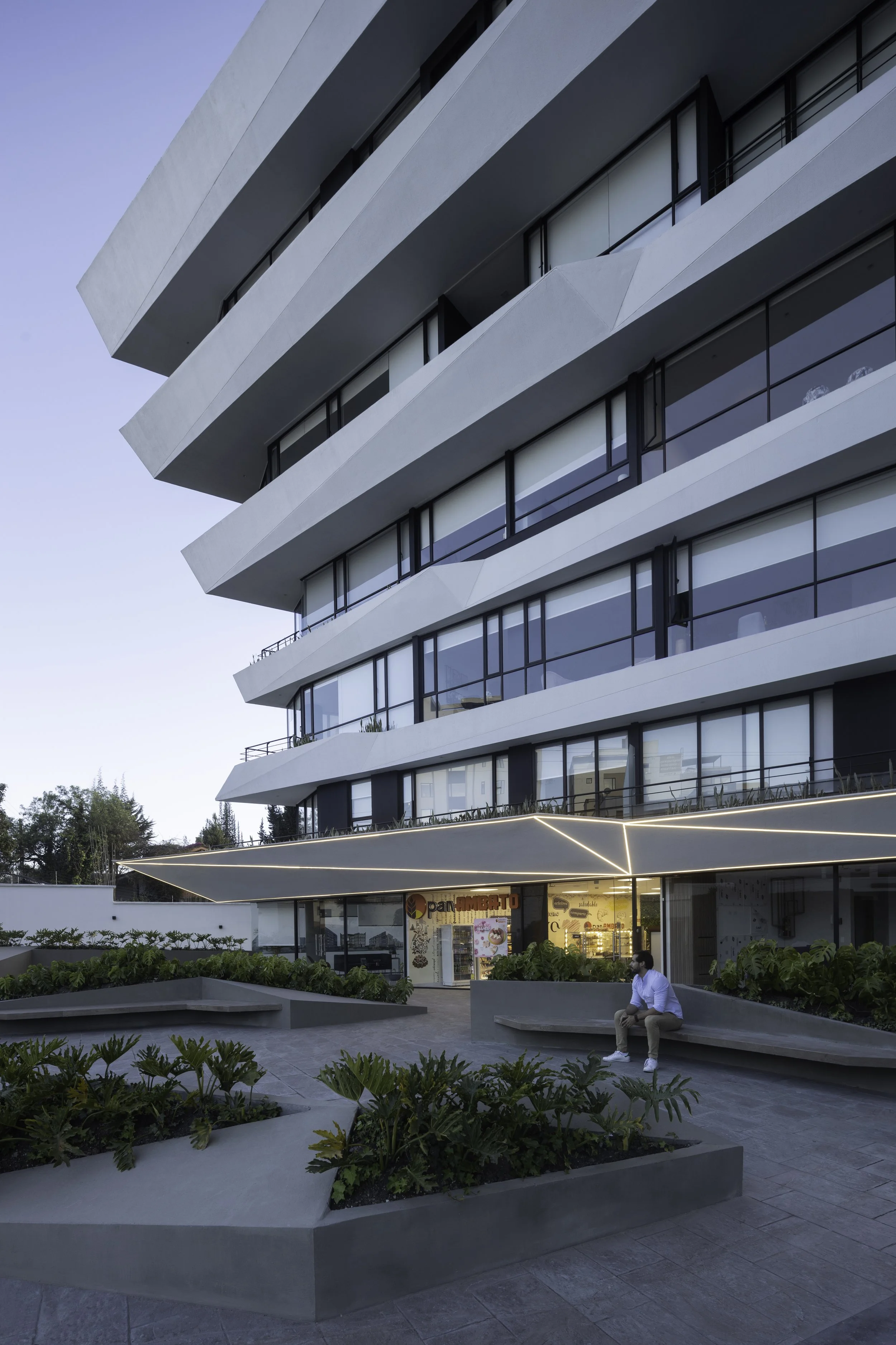 Edificio moderno con balcones, plantas y un hombre sentado en un banco en una plaza frente al edificio.
