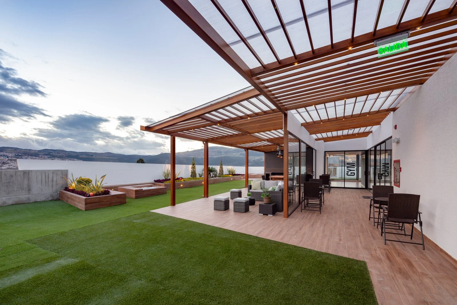 Terraza moderna con piso de madera, área de descanso con sillones y mesas, pérgola de madera, jardín con césped artificial y plantas, muro de concreto, vista hacia una ciudad y colinas, señal de salida en la parte superior.