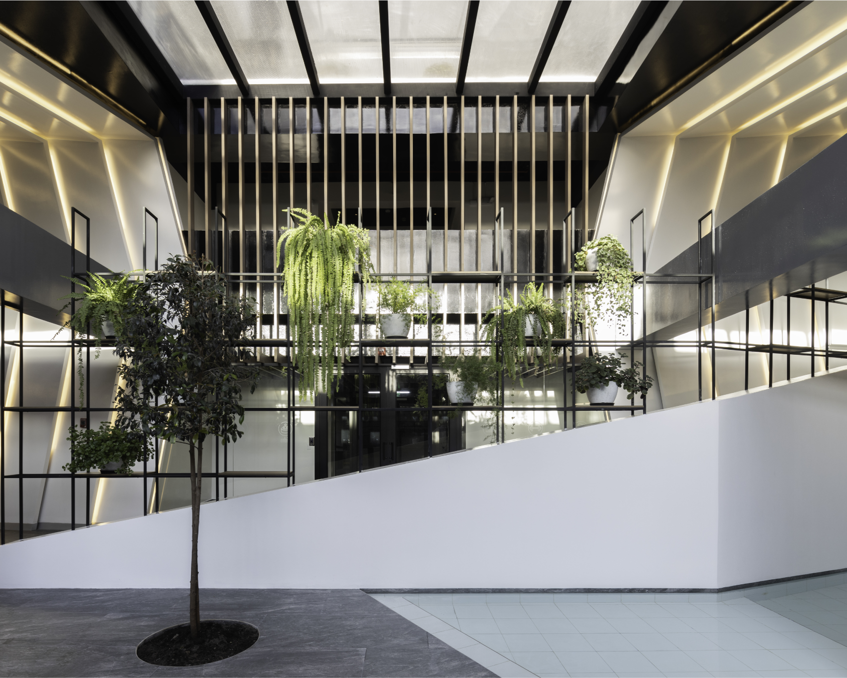 Interior moderno con plantas en macetas blancas y verdes, estructura de marcos negros y paredes blancas con detalles de iluminación en dorado, escalera blanca y piso de piedra y baldosas.