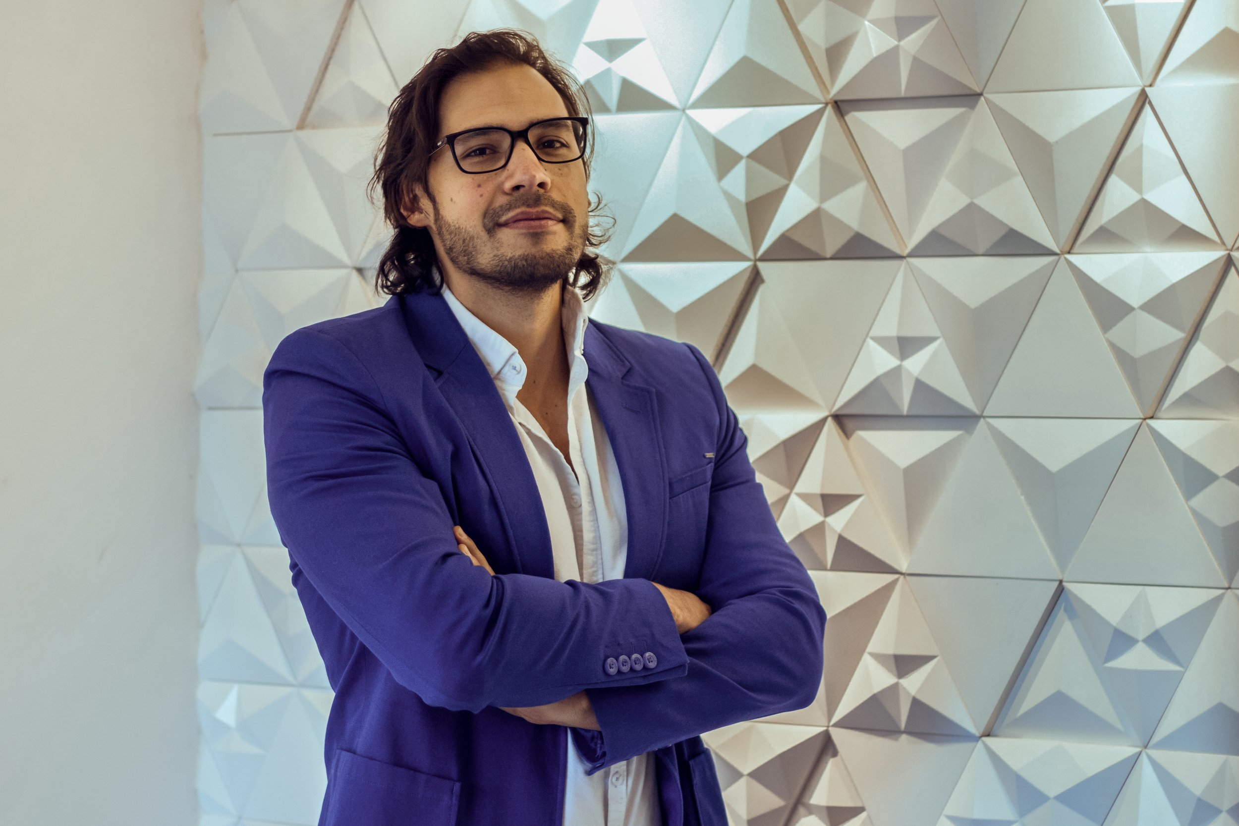 Hombre con gafas y barba, usando saco azul y camisa blanca, con brazos cruzados, de pie frente a pared decorativa de paneles geométricos blancas.