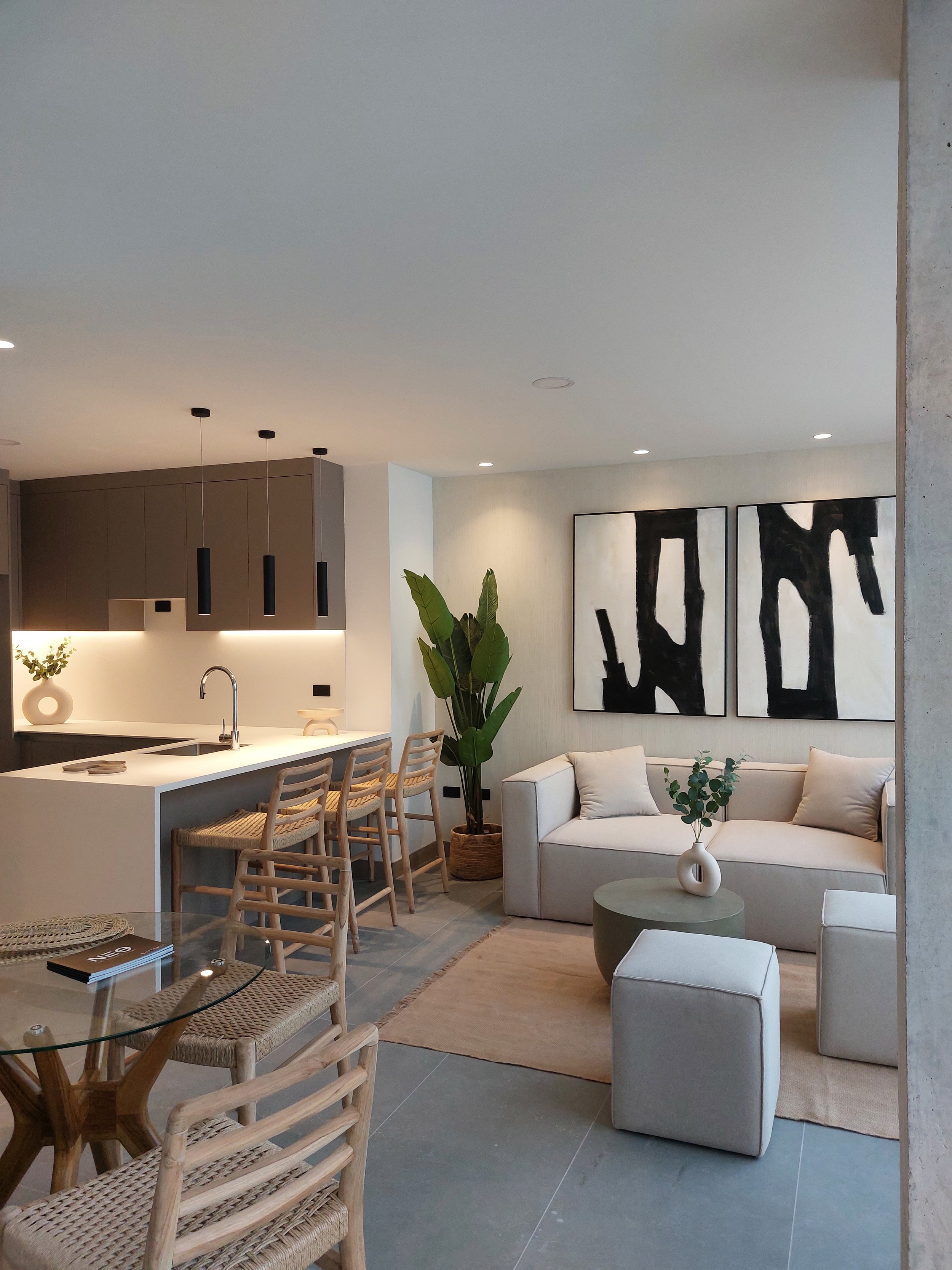 Sala de estar moderna con sofá beige, mesas pequeñas, obras de arte abstractas en la pared, plantas decorativas y una sala de cocina con barra y sillas de madera.