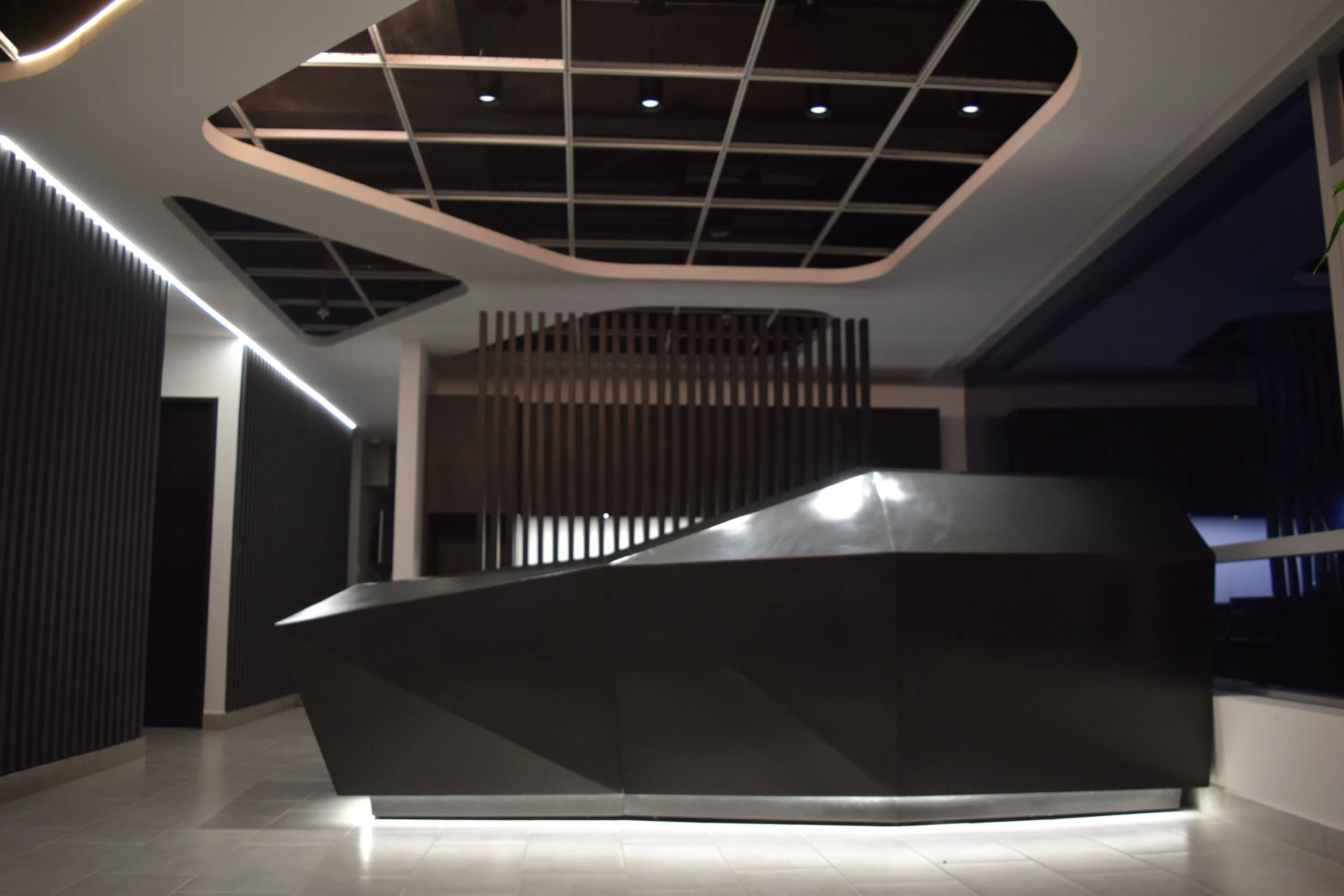 Mostrador de recepción moderno de color gris oscuro en un vestíbulo con paredes negras y techo con detalles de lámparas empotradas.