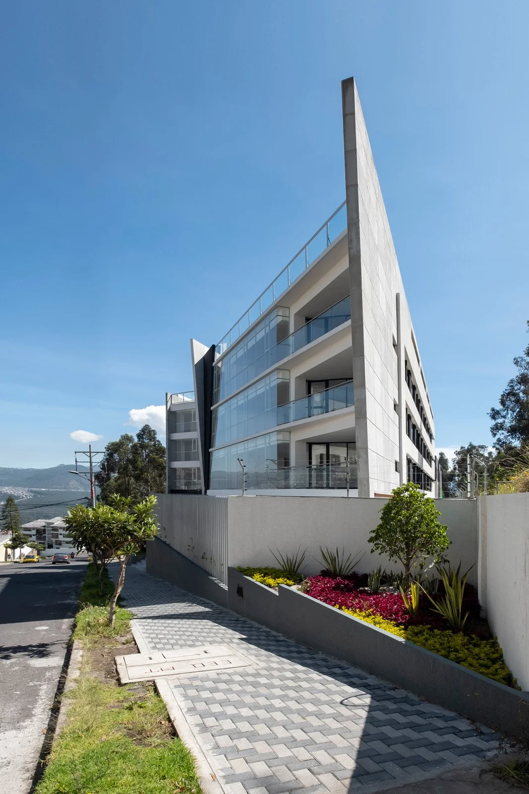 Edificio moderno de varias plantas con balcones de cristal, ubicado en una calle con acera, arboles y vegetación, bajo un cielo azul despejado.