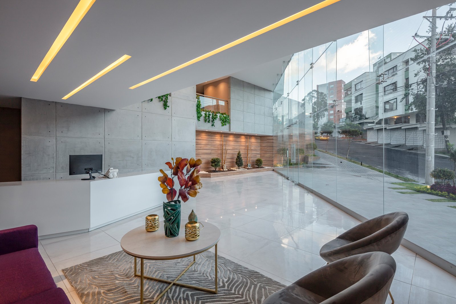 Vestíbulo moderno con recepción blanca, sillones de cuero, mesa decorada con plantas y velas, paredes de concreto y área con plantas y ventanas de vidrio que muestran la calle exterior.