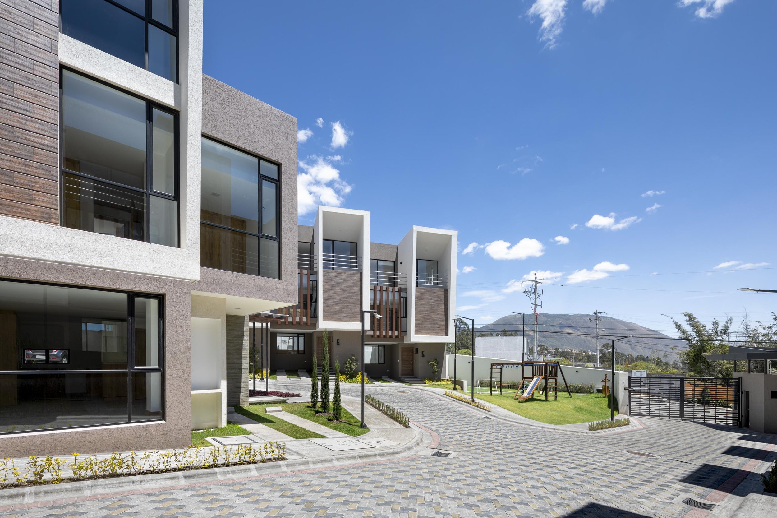 Complejo residencial moderno con departamentos y área recreativa, caminos pavimentados, áreas verdes, juegos infantiles y vistas a montañas en el fondo.