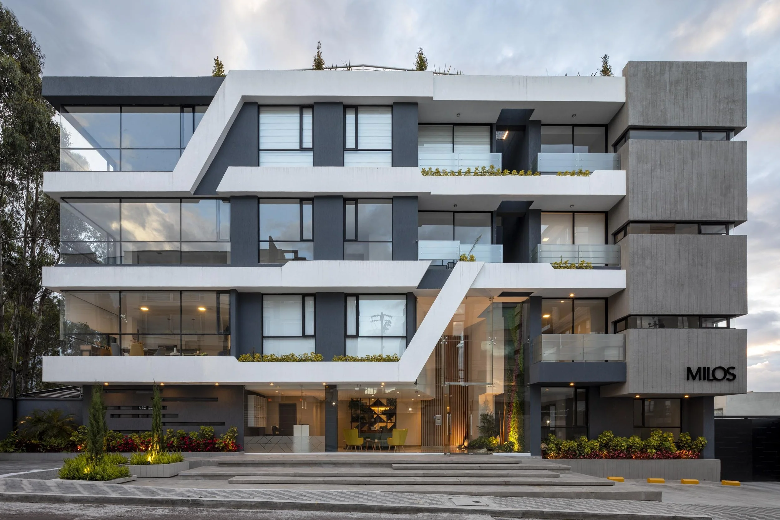 Edificio moderno de varios pisos con balcones y paredes de concreto gris, cartel que dice 'MILOS' en la esquina inferior derecha, plantas y jardineras en la entrada, iluminación cálida en el interior y exterior, y un cielo parcialmente nublado en el 