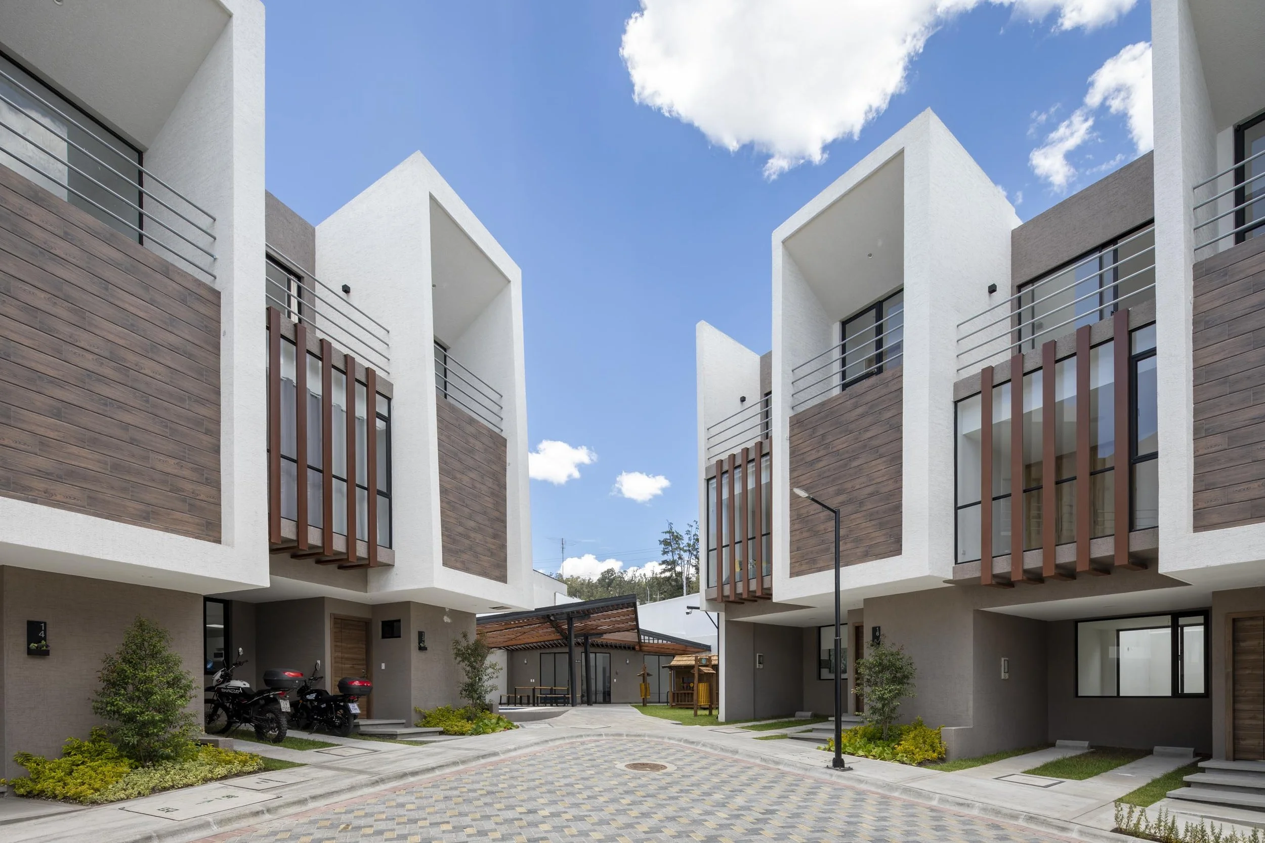 Conjunto de edificios modernos con fachadas de madera y estructura blanca, áreas de estacionamiento con motos y áreas verdes al frente, bajo un cielo azul con nubes
