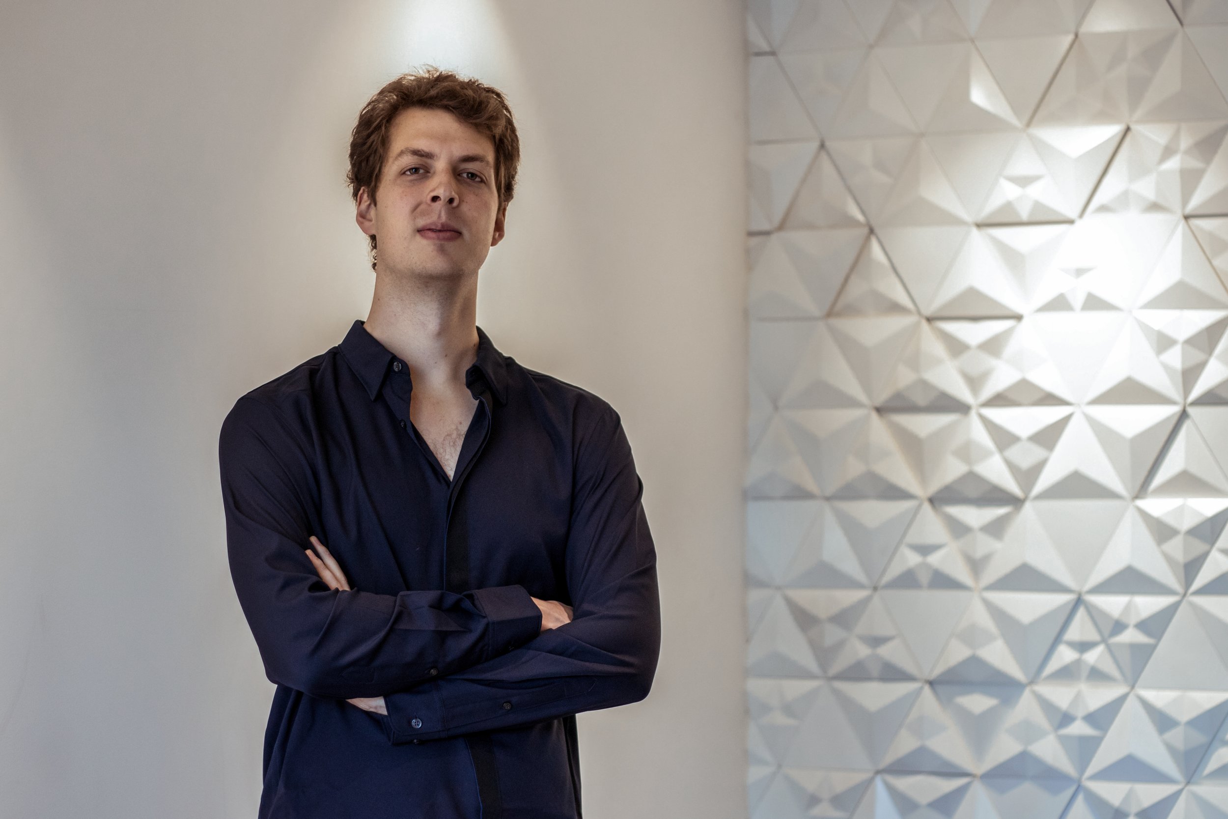 Joven con cabello rizado y camisa negra, de brazos cruzados, en fondo de pared blanca y decorativa.